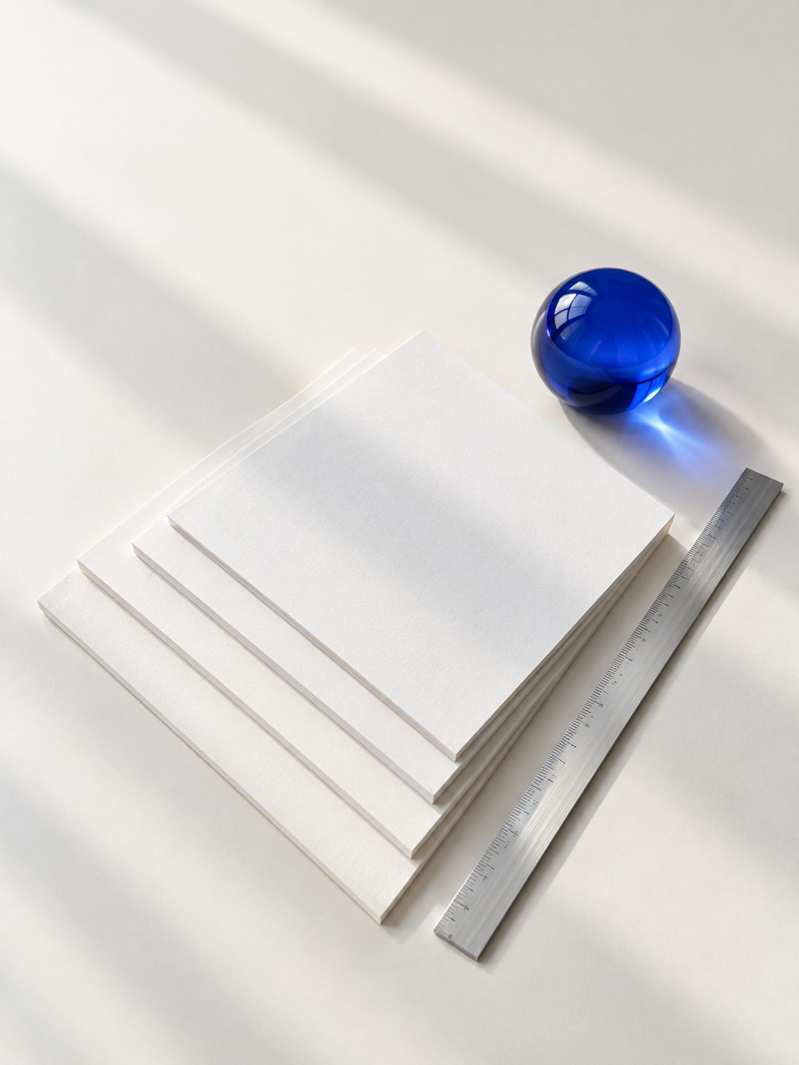 A precise composition of blank white paper sheets layered neatly beside an aluminum rule and a blue glass prism on a bright tabletop.